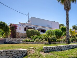 A white building with a tiled roof, stone walls, and a garden with various plants and a palm tree.