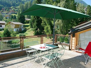 A balcony with a table and chairs, a green umbrella, and a view of a forested hillside. - Bisse-Vieux A-4 by Interhome (Nendaz)