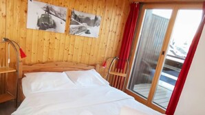 Una habitación de madera con una cama, dos sillas y un ventanal con cortinas rojas.