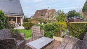 Ein Patio mit Korbmöbeln und einem Tisch, umgeben von Grünpflanzen und Blick auf benachbarte Häuser.