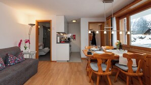 Ein modernes Wohnzimmer mit einer Couch, einem Esstisch mit Stühlen, einer Küchenzeile und Blick auf eine verschneite Landschaft durch große Fenster.