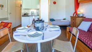 Quarto compacto com uma mesa de jantar posta para quatro, uma cama com colcha xadrez vermelha e branca e uma cozinha compacta ao fundo.