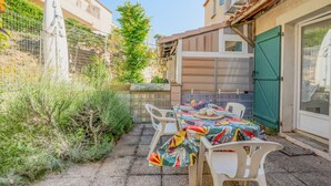 A patio with a table set for two, surrounded by greenery and a building with a tiled roof.