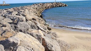 A rocky shoreline with a breakwater extending into the sea, a sandy beach, and a clear sky.