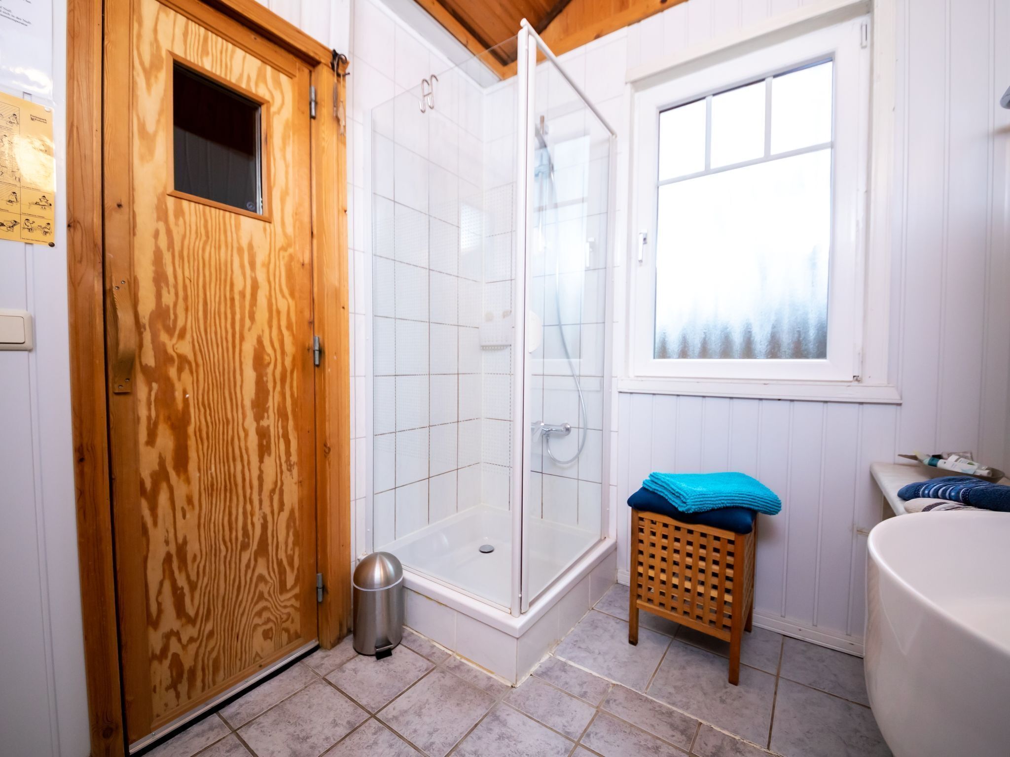 A bathroom with a wooden door, a glass shower enclosure, a small trash bin, a window, and a sink.