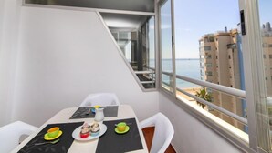 Een moderne kamer met een eettafel gedekt voor twee personen, een raam met uitzicht op zee en gebouwen, en een klein balkon.