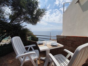 Un balcone con sedie di plastica bianca, un tavolo con piatti e bicchieri, e vista sul mare.