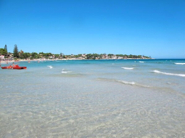 A beach with clear shallow waters, a red boat, and a distant shoreline with buildings.