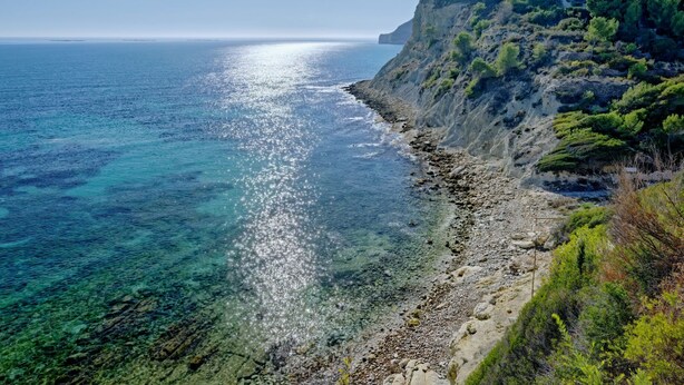 A coastal landscape with a rocky beach, clear turquoise waters, and a cliff with buildings on top. - Apartment Zeus by Interhome (Calpe/Calp)