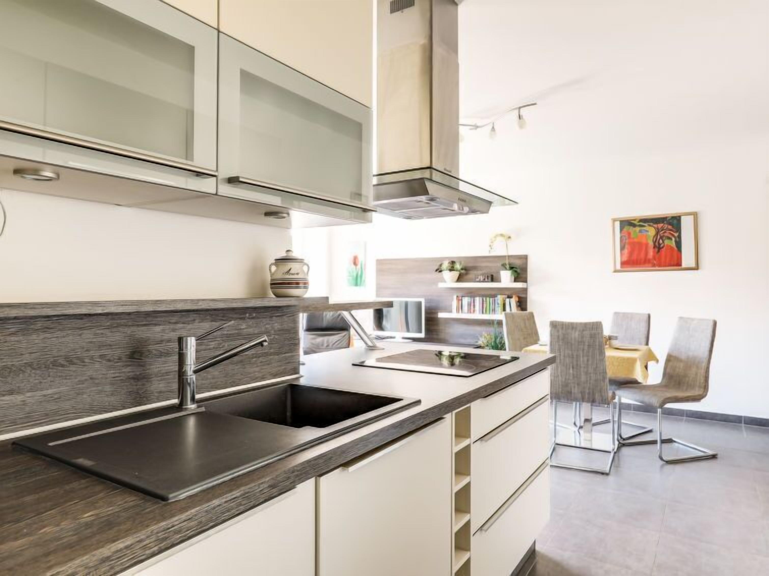 A modern kitchen with a dark wood countertop, stainless steel sink, and white cabinetry. There is a dining area with a table and chairs in the background.