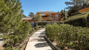 Un chemin menant à un bâtiment doté d’un toit en tuiles, entouré de verdure et d’arbres.