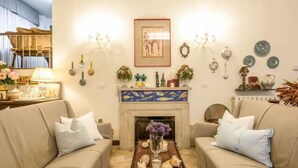 Un salon avec deux canapés beige, une table basse en bois agrémentée d’objets décoratifs, une cheminée aux carreaux colorés et divers éléments de décoration murale, notamment des œuvres d’art encadrées et des appliques murales.