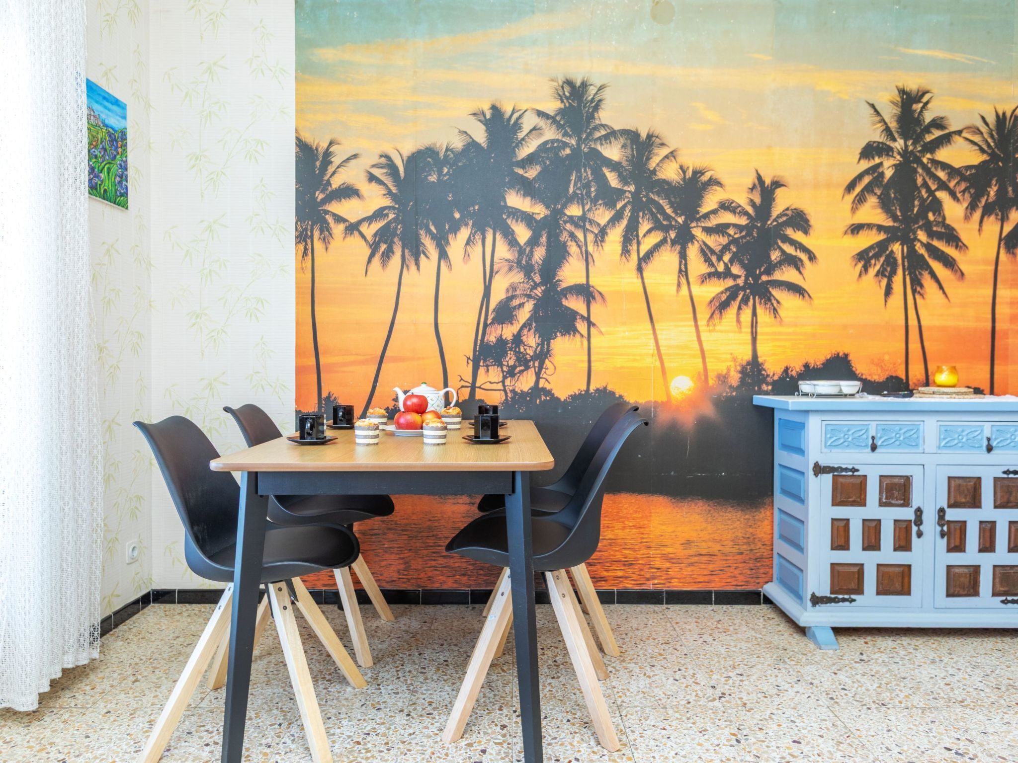 Een eethoek met een tafel voor twee personen, stoelen, een muurschildering van palmbomen en een zonsondergang, en een klein kastje met lades.