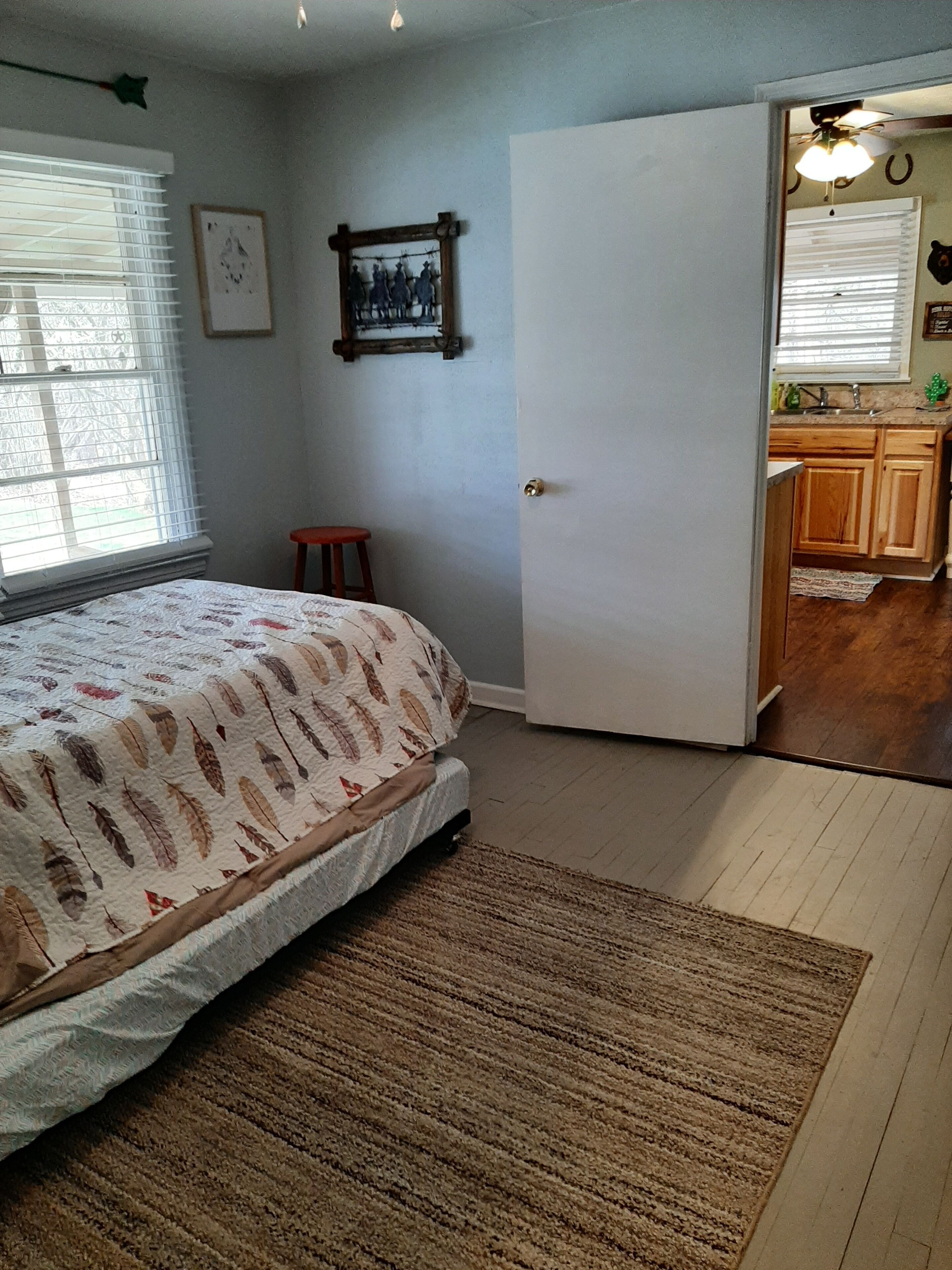 A bedroom with a bed, a small stool, a door leading to a kitchen, and a window with blinds.