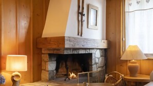 Una stanza accogliente con camino in pietra, pareti in legno, una zona salotto con cuscini e una lampada su un tavolino.