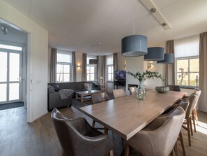 Un salon moderne avec un canapé, une table basse et un téléviseur, un coin repas avec une table et des chaises en bois, et de grandes fenêtres avec des rideaux.
