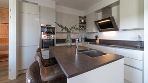 Una cocina moderna con armarios blancos, electrodomésticos de acero inoxidable, una isla central con un lavamanos y una zona de estar con un banco.
