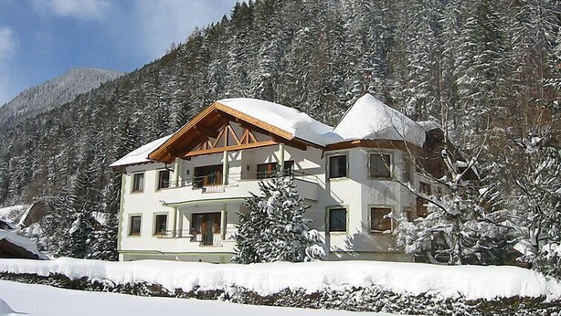 Eine zweigeschossige Chalet mit geneigtem Dach, umgeben von schneebedeckten Bäumen und einem Berg im Hintergrund.