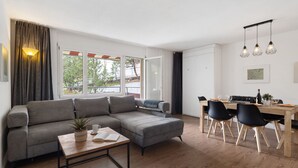 A modern living room with a grey sectional sofa, a wooden coffee table, and a dining area with a wooden table and black chairs. - Allod Park Haus C 105 by Interhome (Davos)