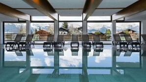 Ein großer, überdachter Pool mit Panoramafenstern, mehreren Liegestühlen und Blick auf Berge und Gebäude.
