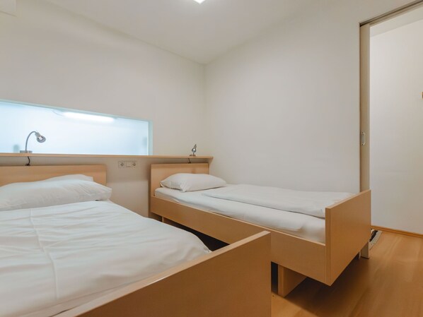 A room with two single beds, each with a wooden headboard and white bedding. There is a window with a light fixture above the left bed, and a door on the right.