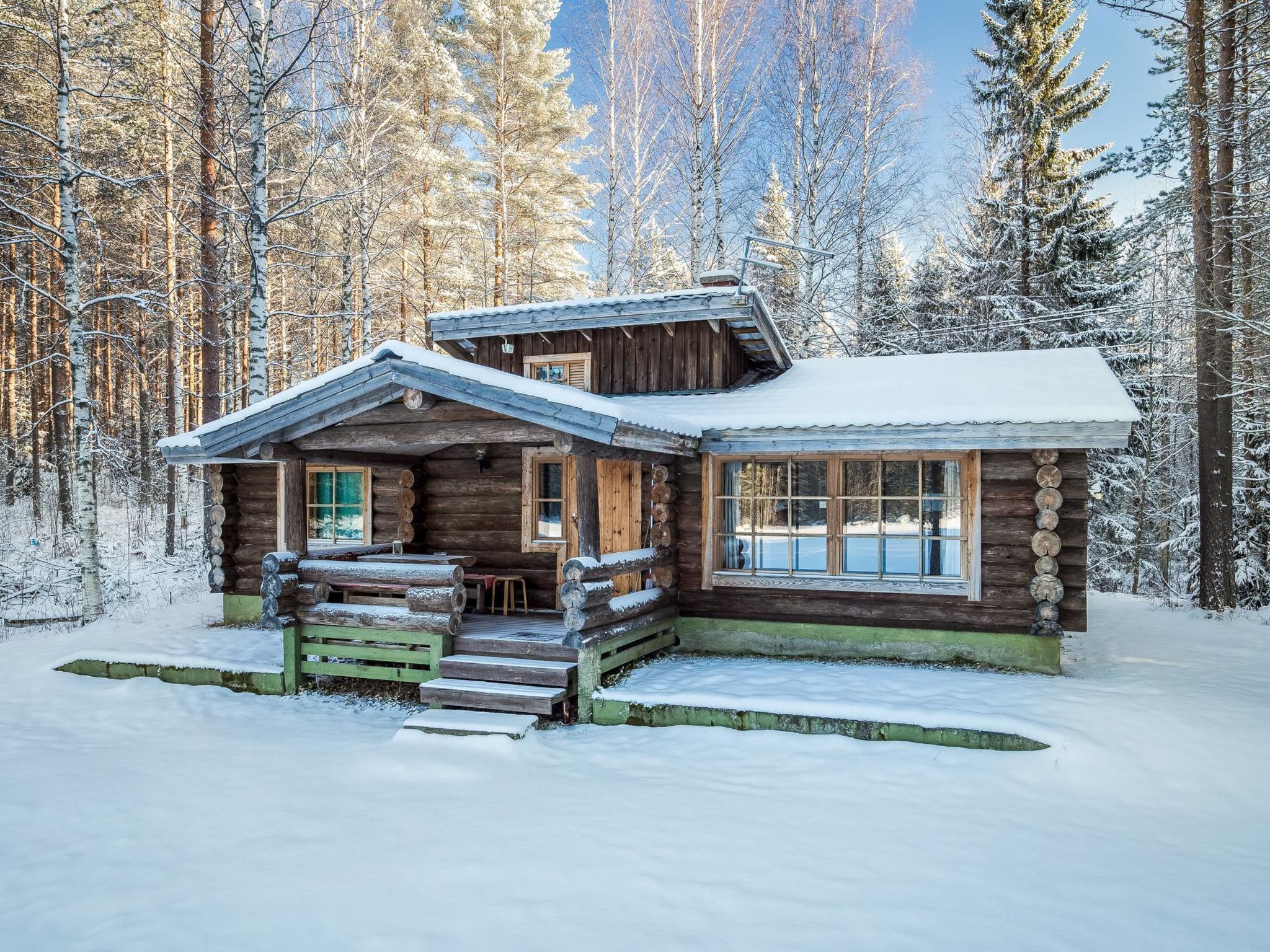 A log cabin with a porch and a window, surrounded by snow-covered trees.