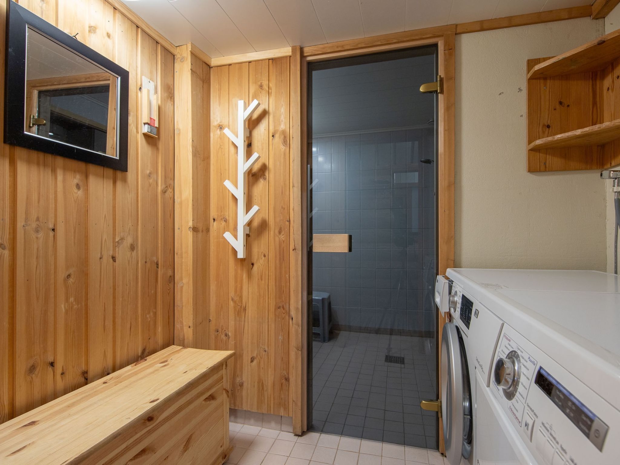 Un interior de madera con un banco, un espejo, una lavadora blanca y una puerta que da a un cuarto con azulejos.