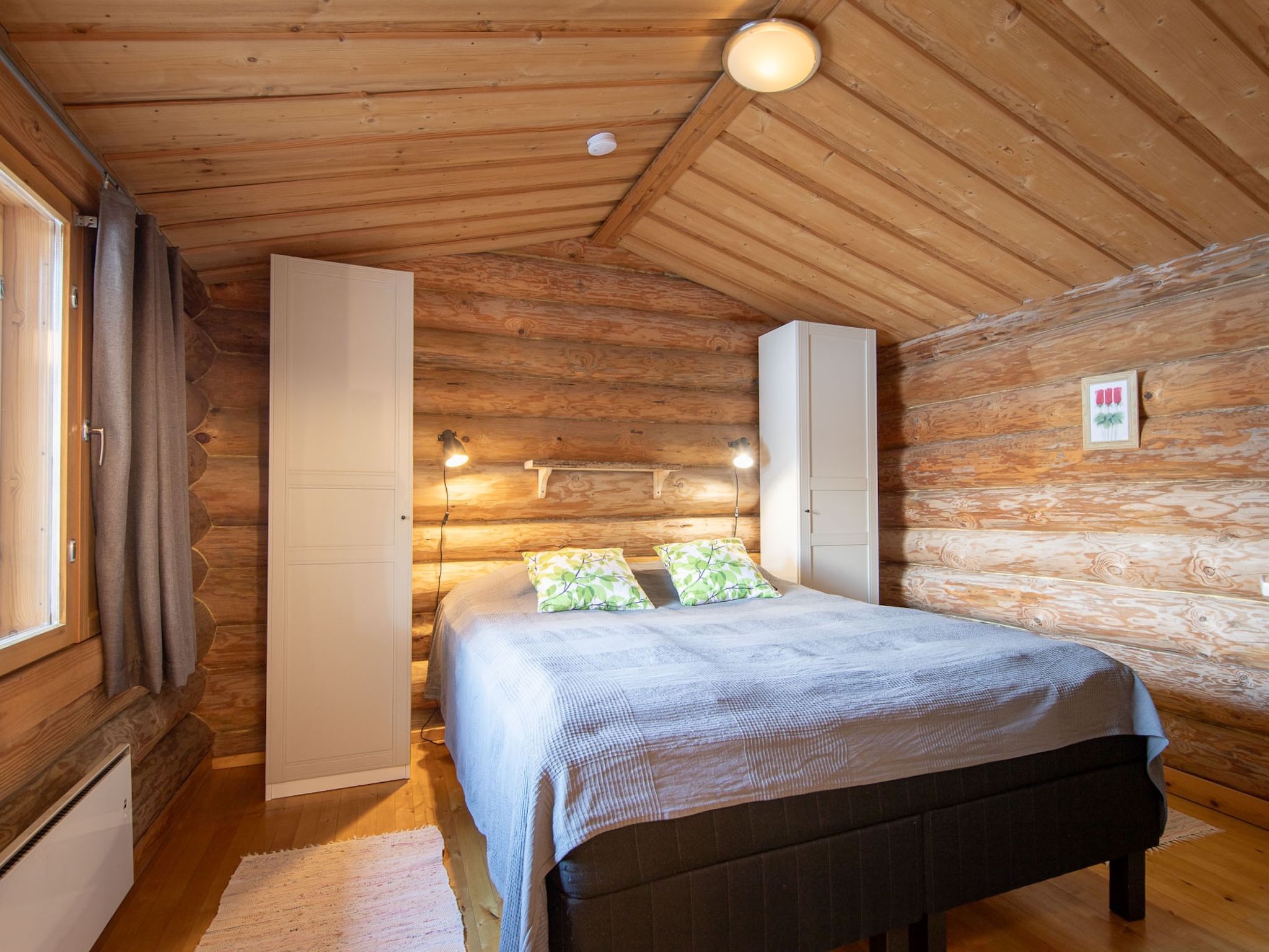 A log cabin bedroom with a bed, two white wardrobes, a window with curtains, and a wall-mounted light fixture.