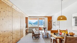 Um quarto acolhedor com um guarda-roupa de madeira, um sofá, uma mesa de jantar com cadeiras e vista para montanhas através de uma janela grande.
