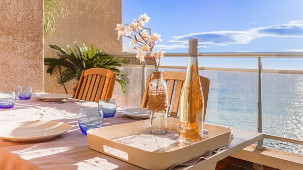 Een gedekte tafel met borden, glazen en een fles op een balkon met uitzicht op zee.