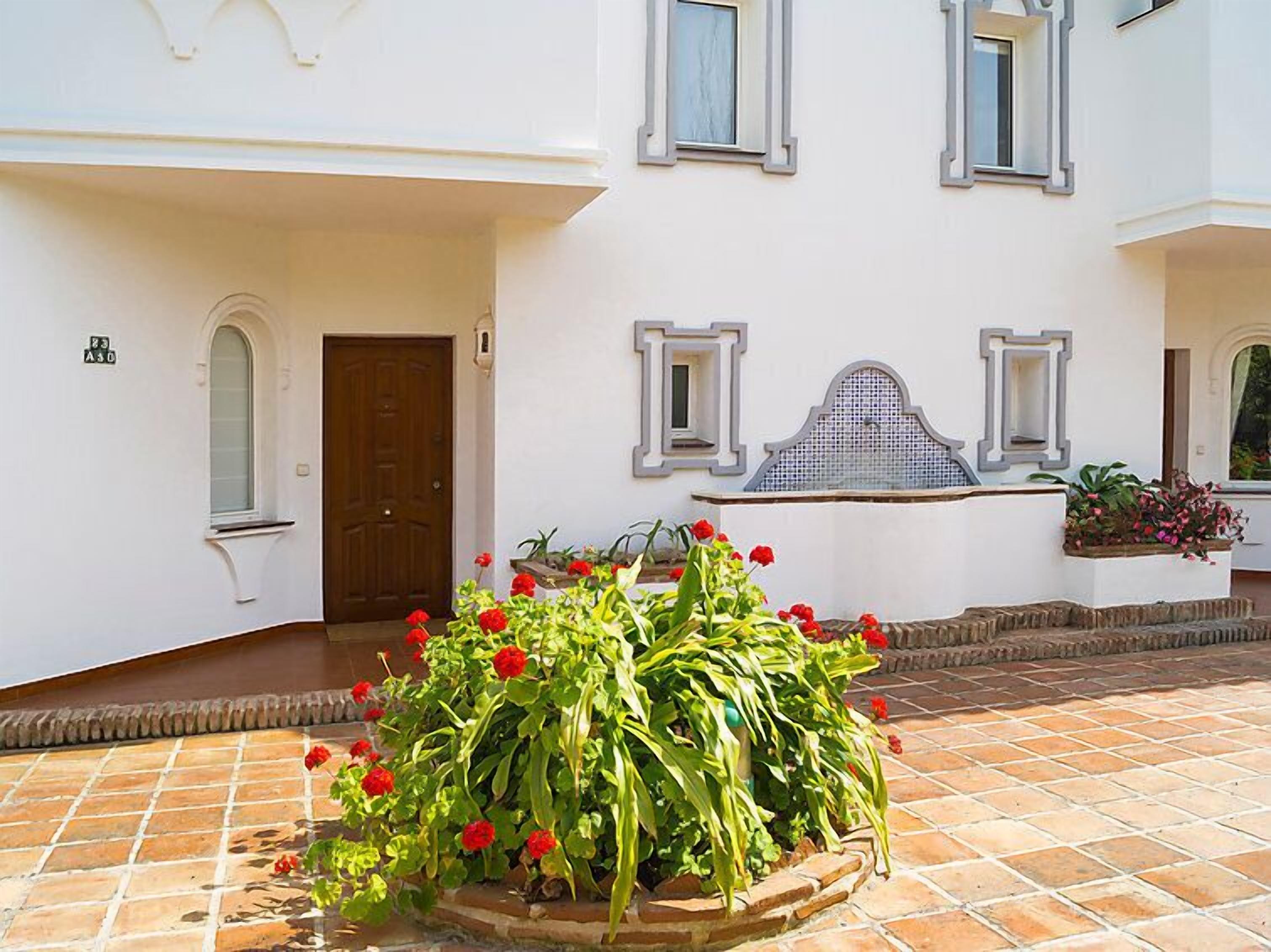 Ein weißes Gebäude mit einer mit Fliesen belegten Terrasse, einer Holztür und einem Blumenbeet mit roten Blüten.