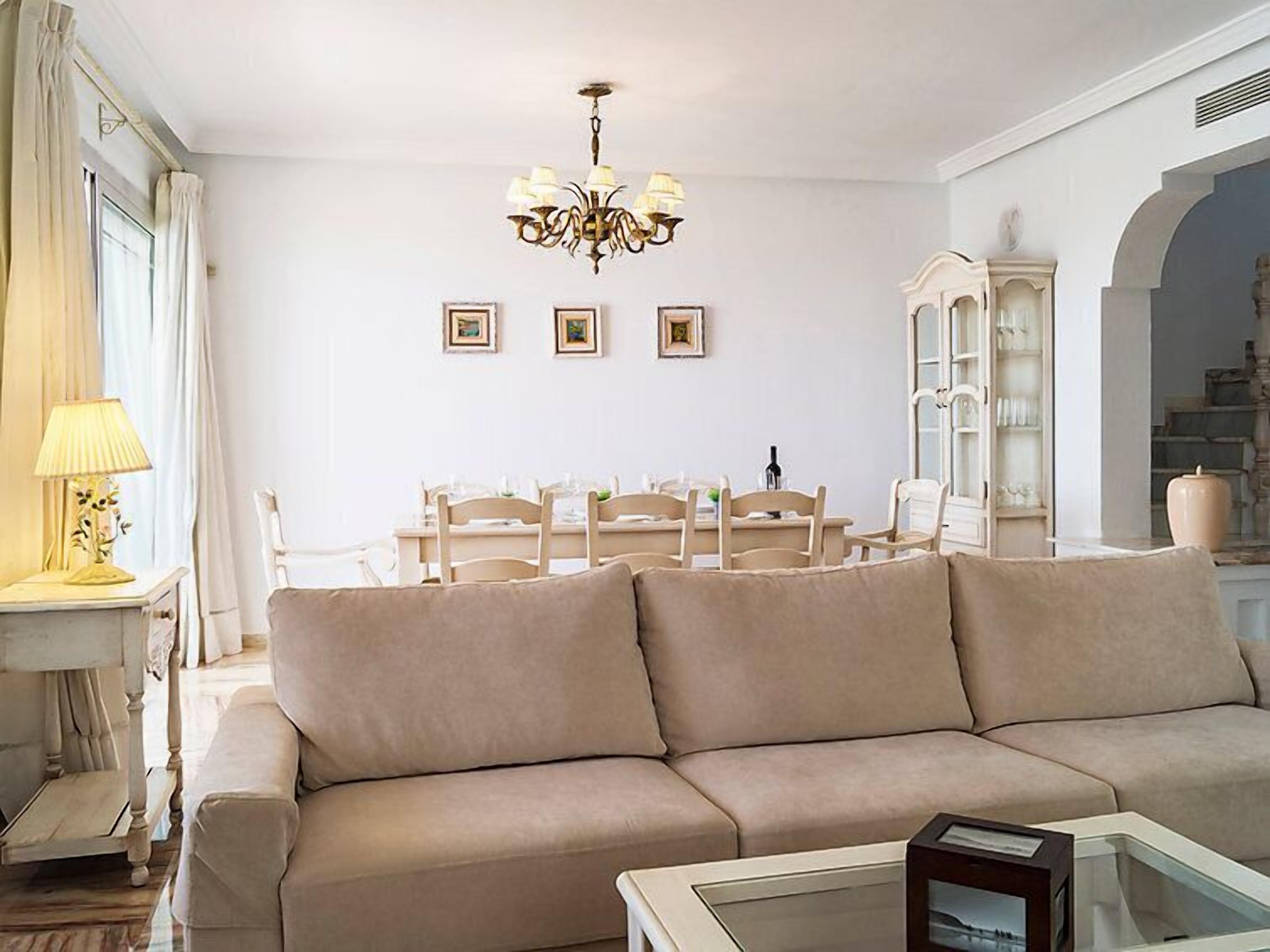 A living room with a beige sofa, a glass coffee table, a chandelier, and a dining area with a white table and chairs.