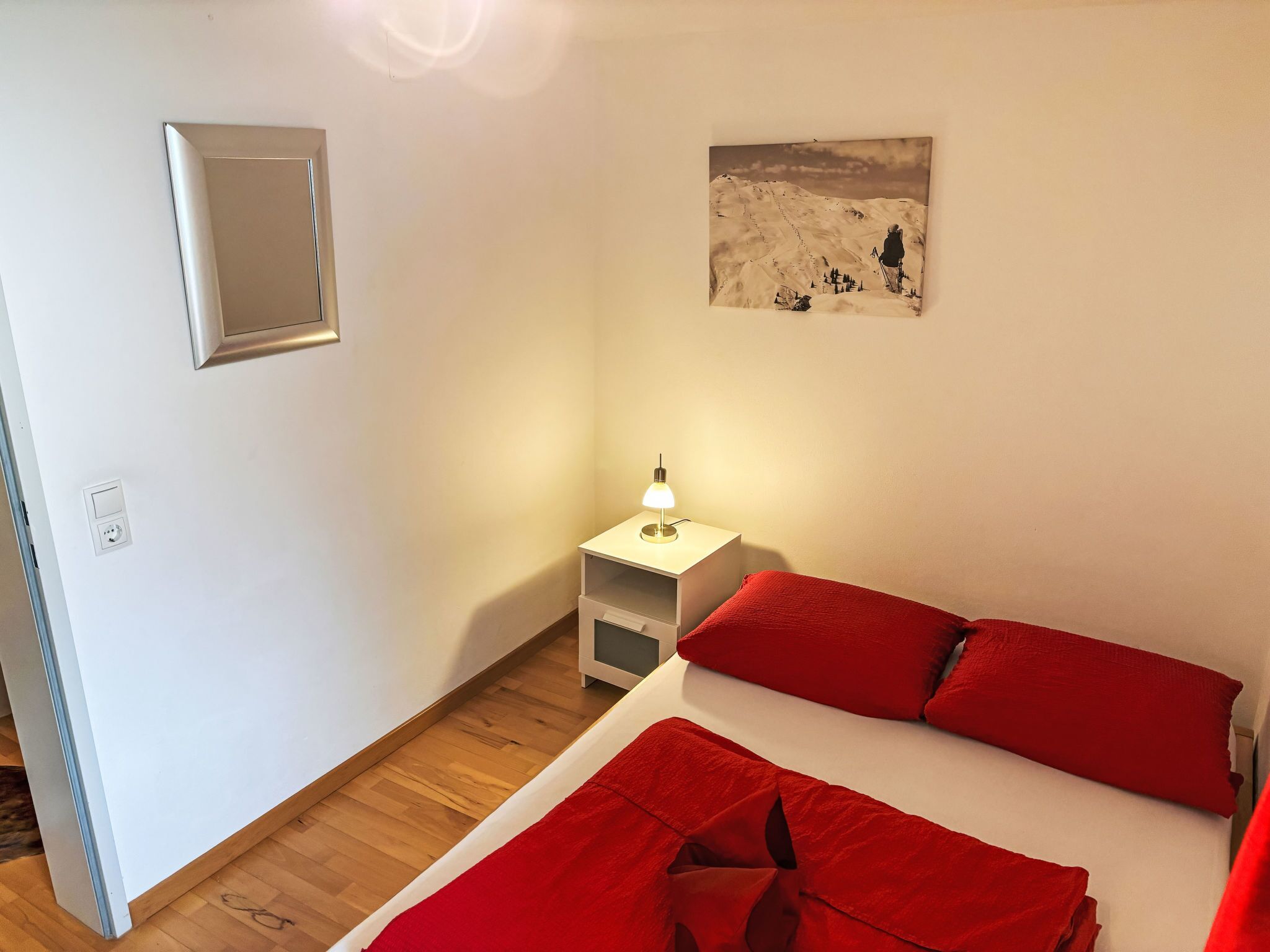 Een slaapkamer met een bed, rode kussens, een nachtkastje met een lamp, een schilderij aan de muur en een klein raam.
