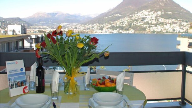 Una mesa para dos, con un jarrón de flores, una botella de vino y un frutero. Se aprecia una vista de una montaña y un cuerpo de agua al fondo.