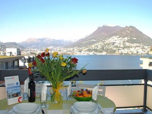Une table dressée pour deux personnes, avec un vase de fleurs, une bouteille de vin et un saladier de fruits. On aperçoit en arrière-plan une montagne et une étendue d'eau.