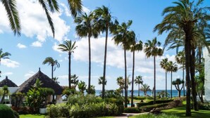Um resort tropical com um edifício de telhado de palha, palmeiras, um gramado bem cuidado e um caminho que leva a uma área de piscina.