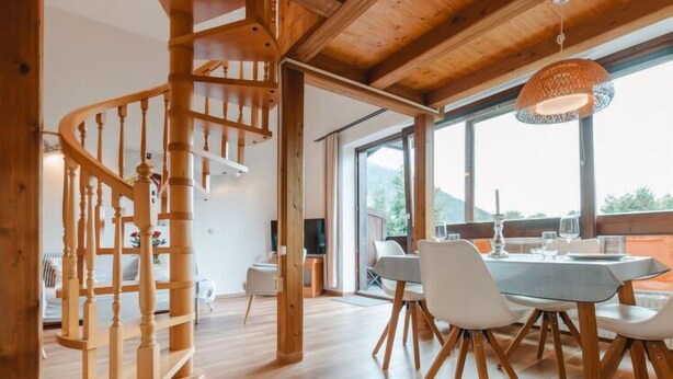 A modern interior with a wooden spiral staircase, a dining area with a round table and chairs, and large windows offering a view of the outdoors. - Anna-Katharina by Interhome (Bad Hofgastein)