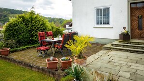 Une maison blanche avec une porte en bois, une terrasse en briques et une table avec des chaises.