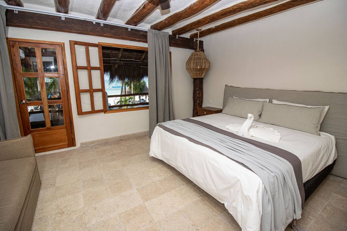 Un dormitorio con una cama grande, una puerta de madera, una ventana con vistas a palmeras y una lámpara colgante.
