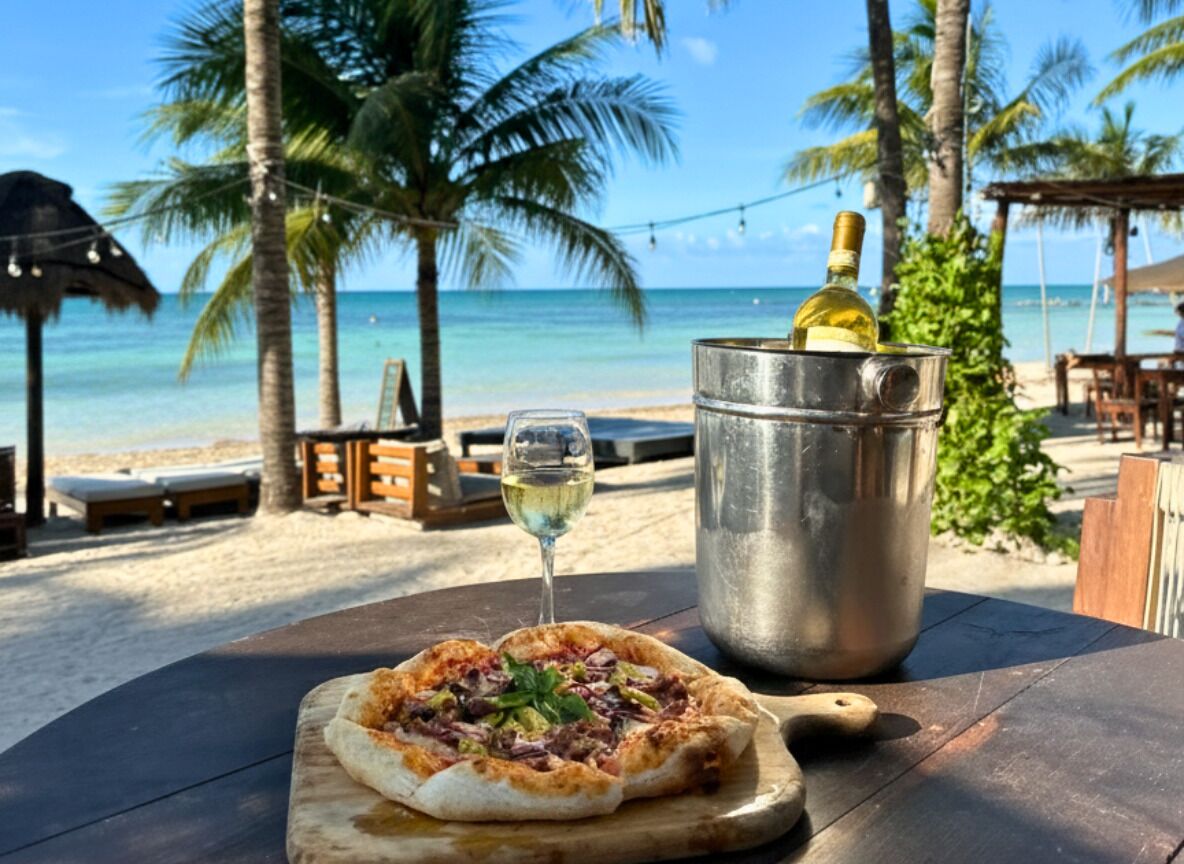 Una pizza sobre una tabla de madera, una copa de vino y una botella de vino en un balde de metal sobre una mesa en la playa, con palmeras y el océano al fondo.