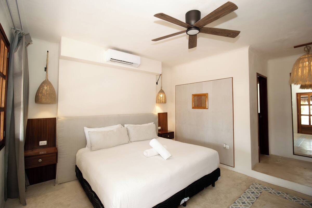 Un dormitorio con cama, ventilador de techo, unidad de aire acondicionado, mesita de noche, lámpara y lámpara colgante.