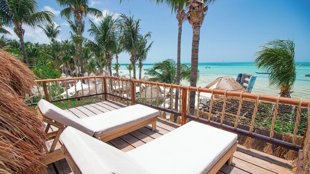 A wooden deck with two lounge chairs, a thatched structure, and a view of a beach with palm trees and a clear sky. - Amaité Beach Hotel (Isla Holbox)