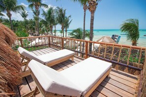 A wooden deck with two lounge chairs, a thatched structure, and a view of a beach with palm trees and a clear sky. - Amaité Beach Hotel (Isla Holbox)