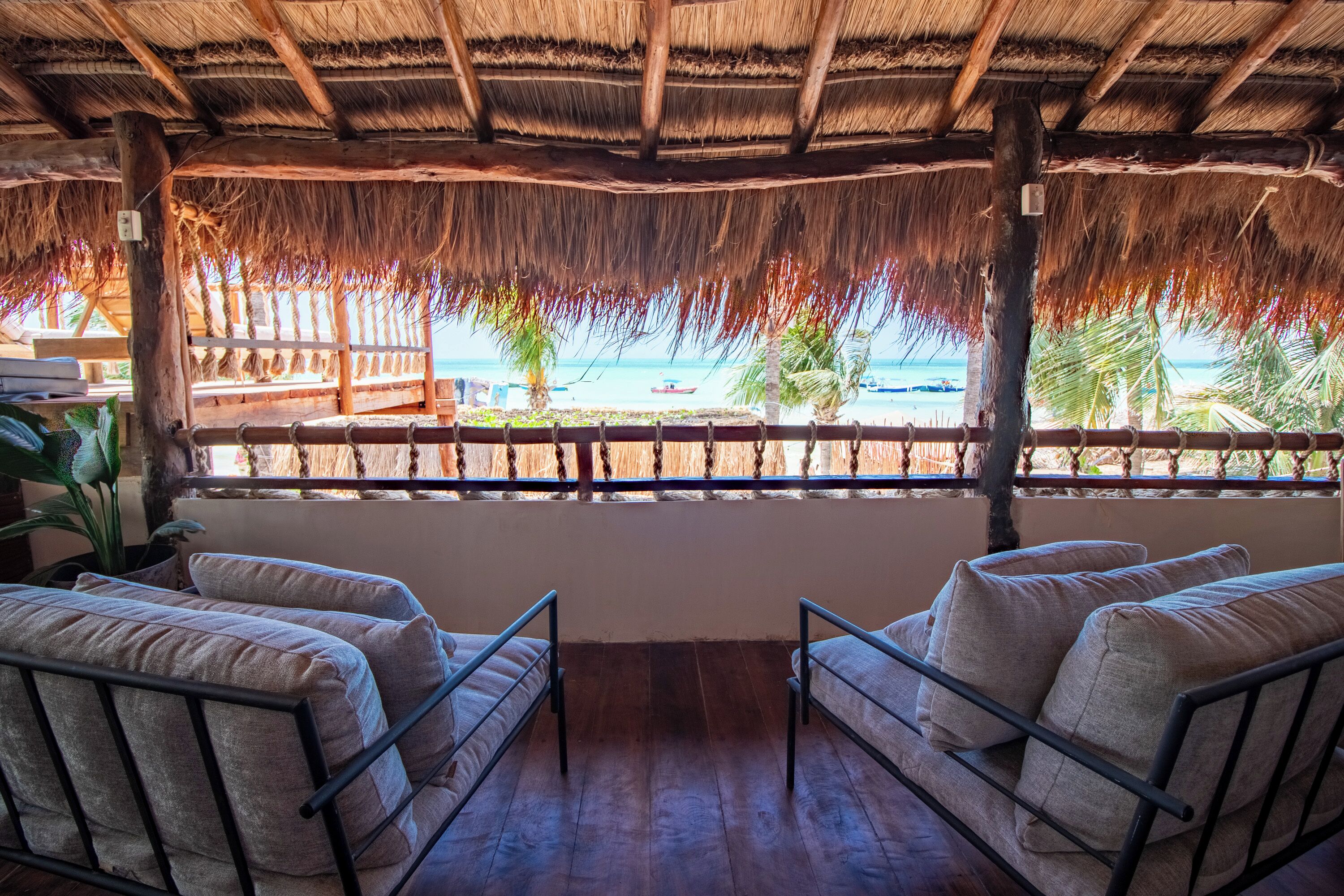 A room with a thatched roof, wooden floor, and two cushioned chairs facing a beach view.