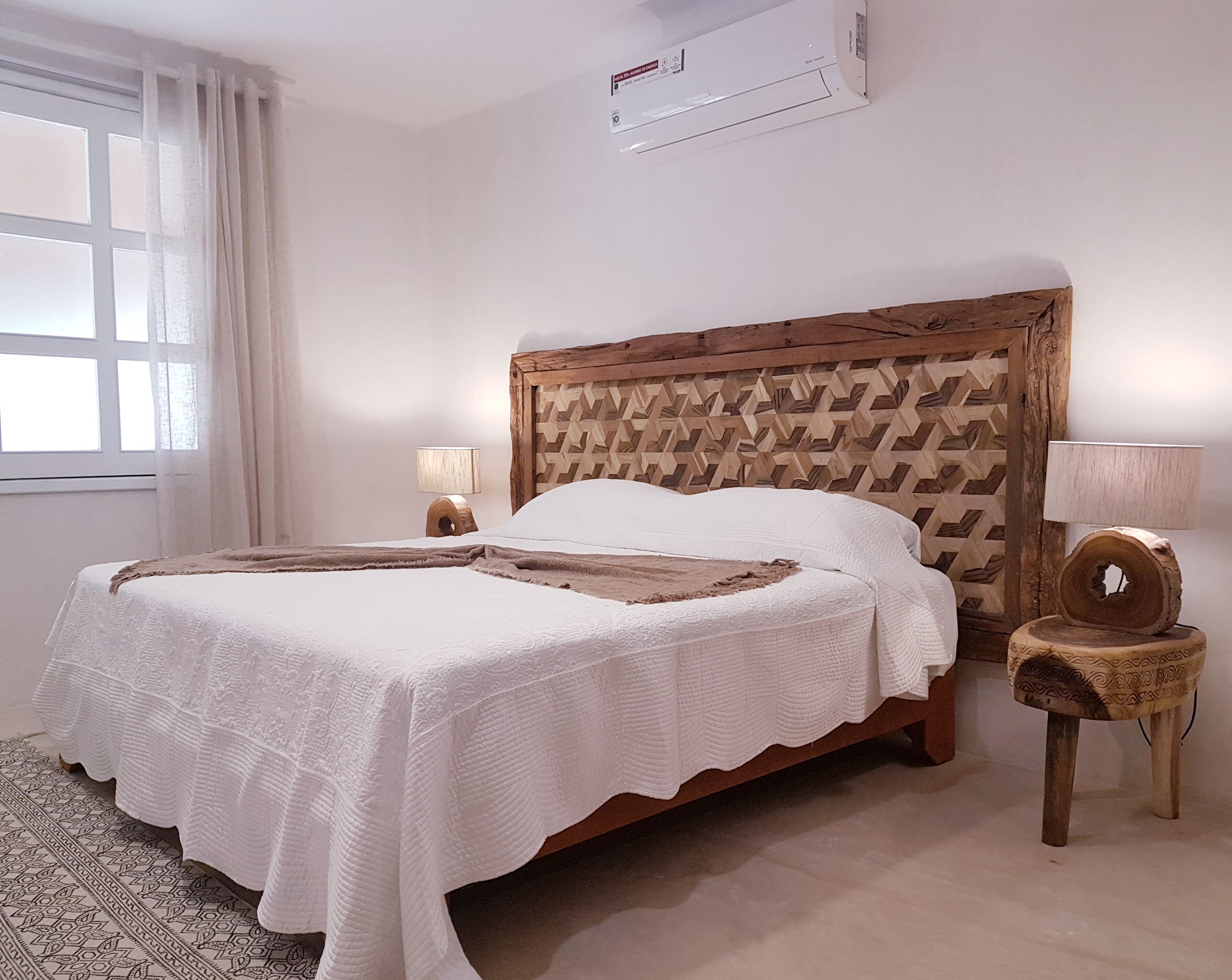 Une chambre à coucher avec une tête de lit en bois, un lit recouvert de draps blancs, une lampe de chevet et une fenêtre avec des rideaux.