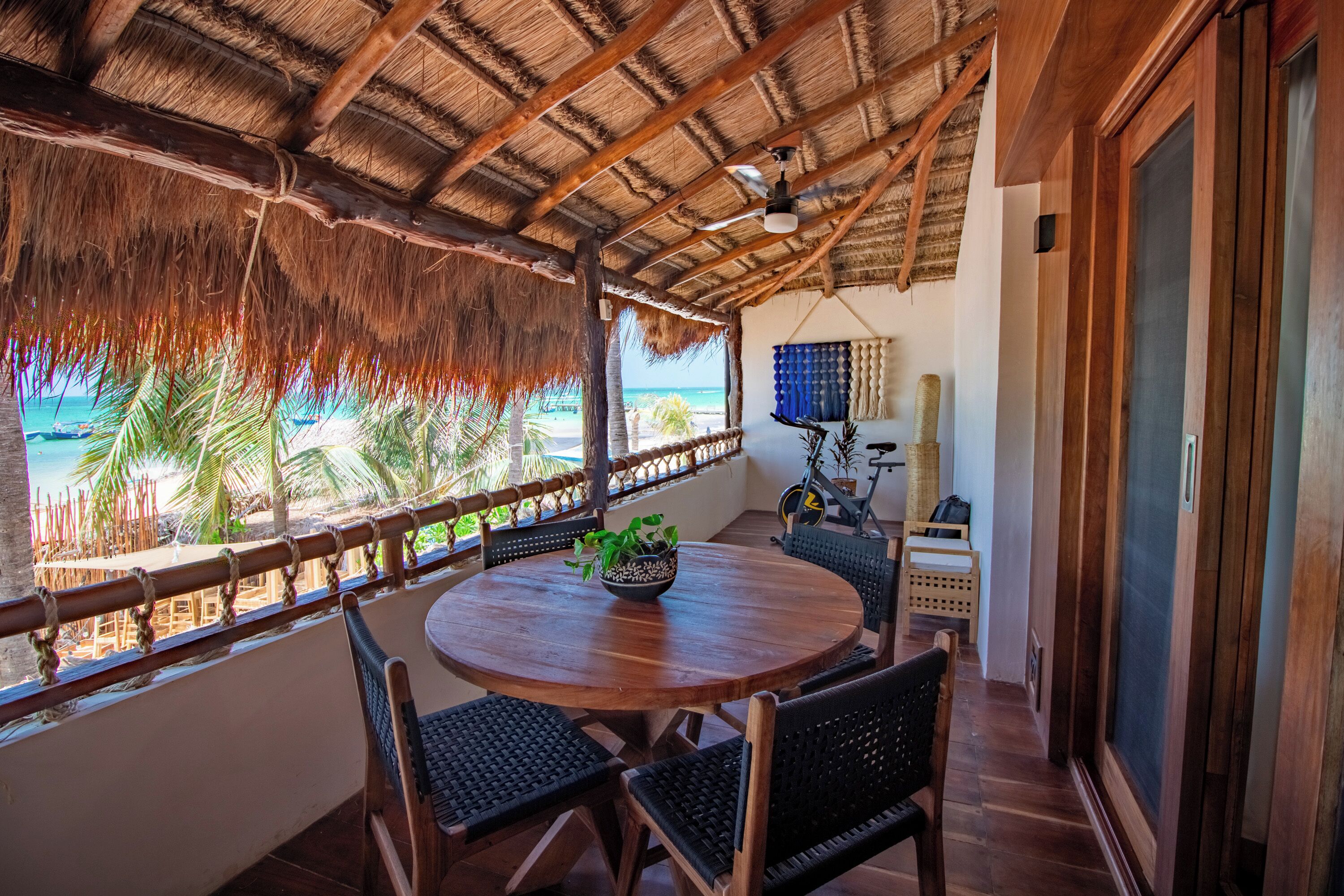 A wooden table with chairs on a balcony overlooking the ocean, with a thatched roof and wooden railings.