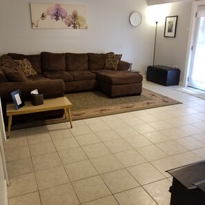 A living room with a brown sectional sofa, a small wooden table, a clock on the wall, and a rug on the floor. - Private Basement Apartment (Athens)