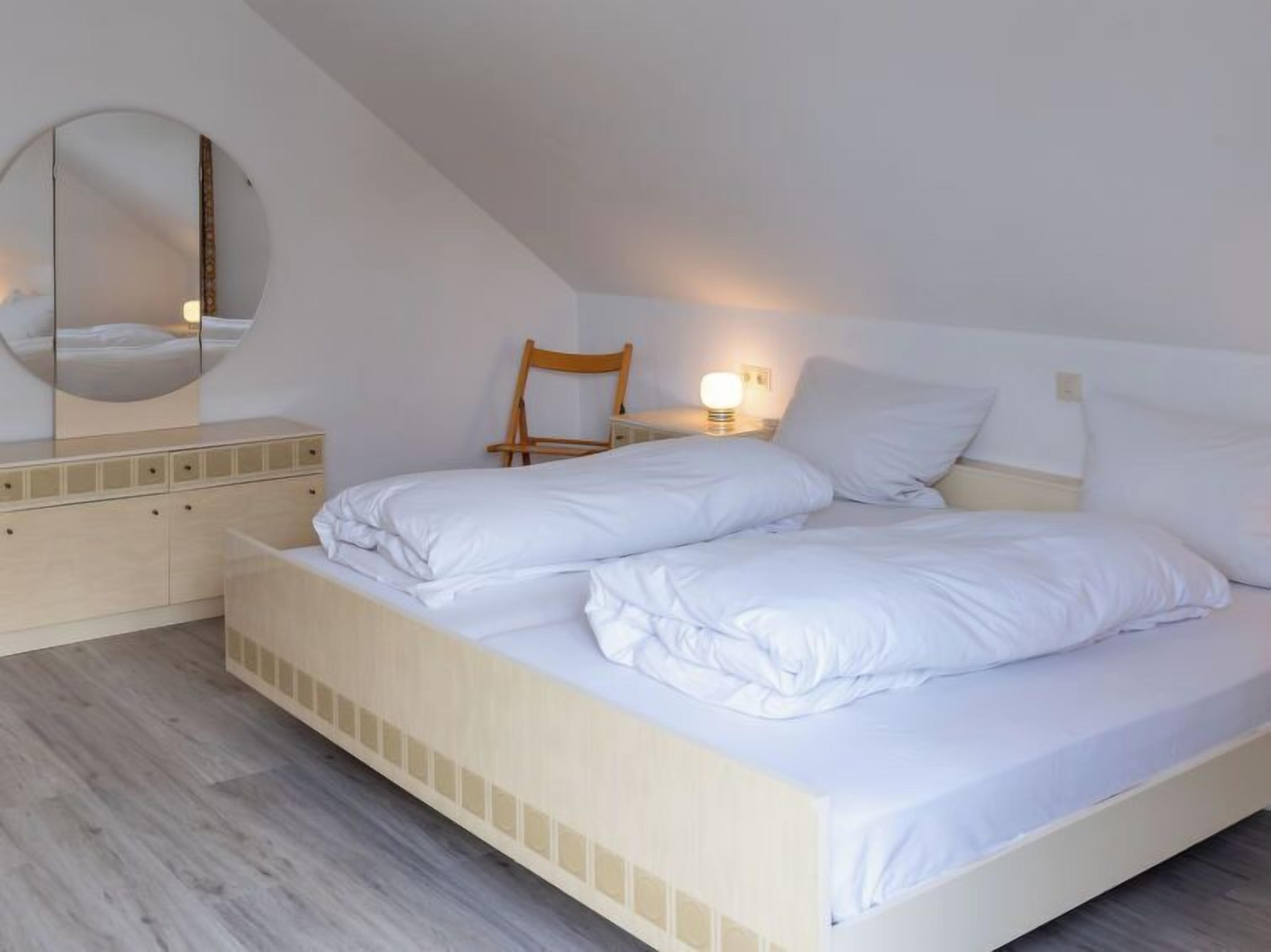 Un dormitorio con cama, una silla de madera, una mesita de noche con lámpara y un espejo en la pared.