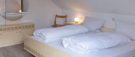 Un dormitorio con cama, una silla de madera, una mesita de noche con lámpara y un espejo en la pared.