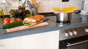 Una cocina con tabla de cortar, verduras, un pan y una olla de espagueti sobre la encimera.